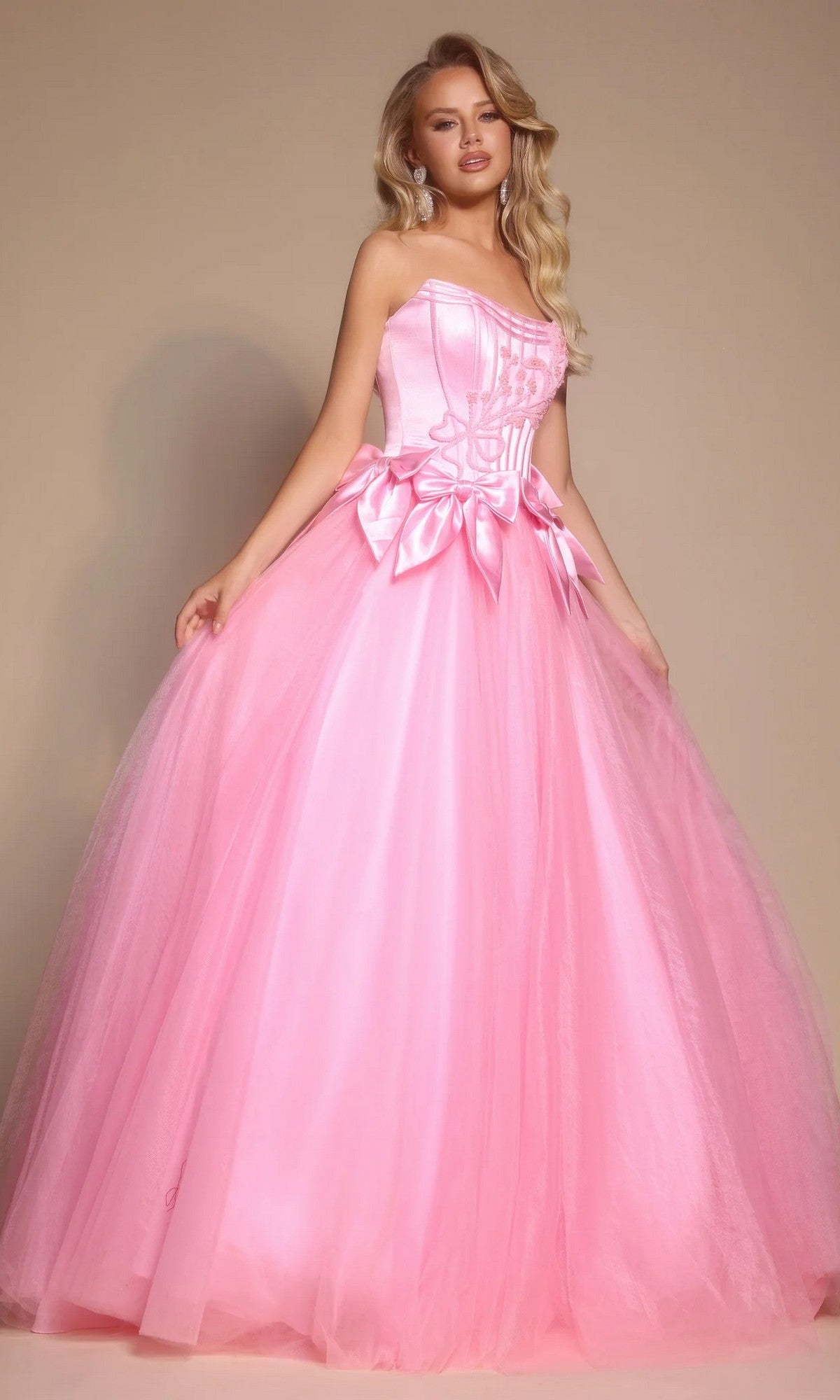 A woman stands against a neutral background wearing the Portia and Scarlett PS26102 strapless ball gown in light pink, featuring a corset bodice and full tulle skirt.